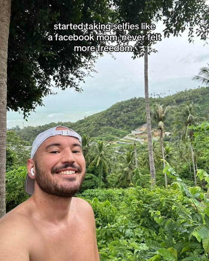 Smiling man taking a selfie outdoors with lush green trees and hills, capturing the chaotic energy of a mom on social media.