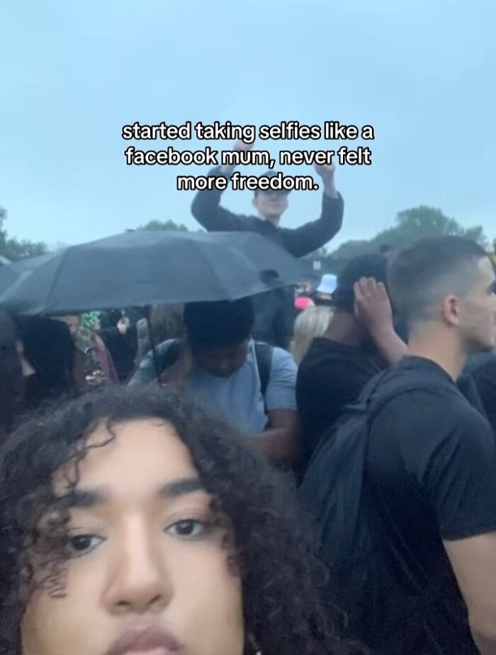 Woman with curly hair taking a selfie in a crowded outdoor setting, capturing chaotic energy of a mom on social media.