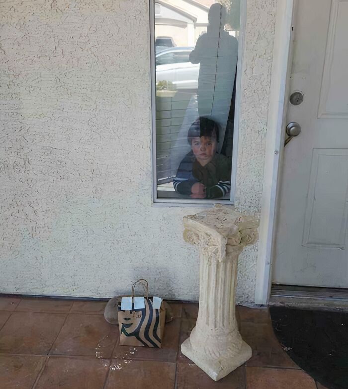 Delivery confirmation pic shows a package left beside a broken pillar with a child watching through a window.