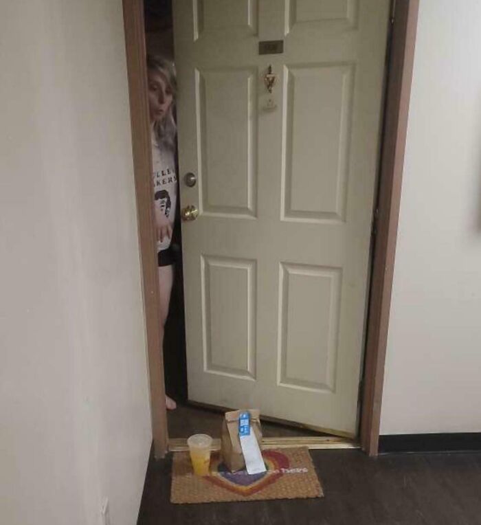Delivery confirmation pic showing a food order left at the door with a customer peeking out behind it.