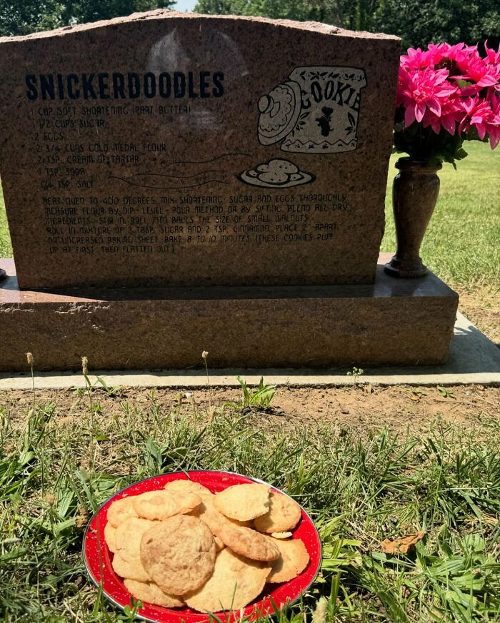 Tombstone etched with snickerdoodles recipe beside a red plate of cookies and pink flowers in a cemetery setting.