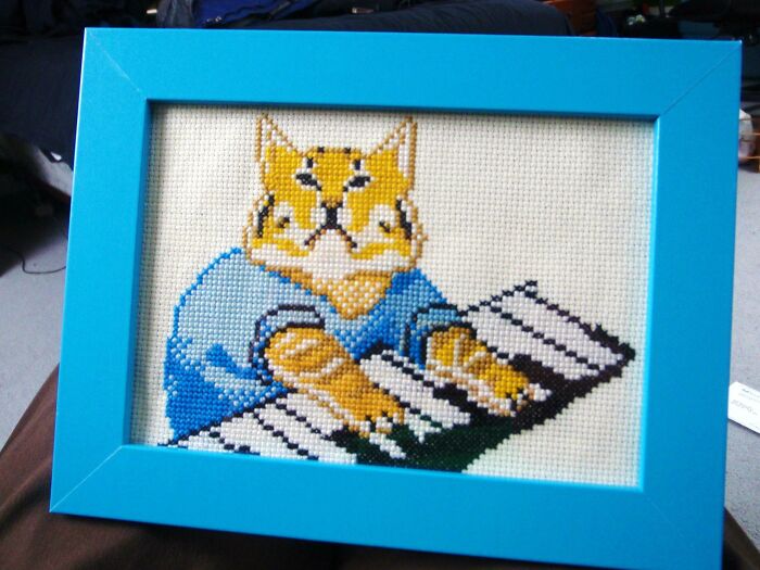 Cross-stitched cat playing piano in blue frame, a funny Valentine’s Day gift showing love with humor and creativity.