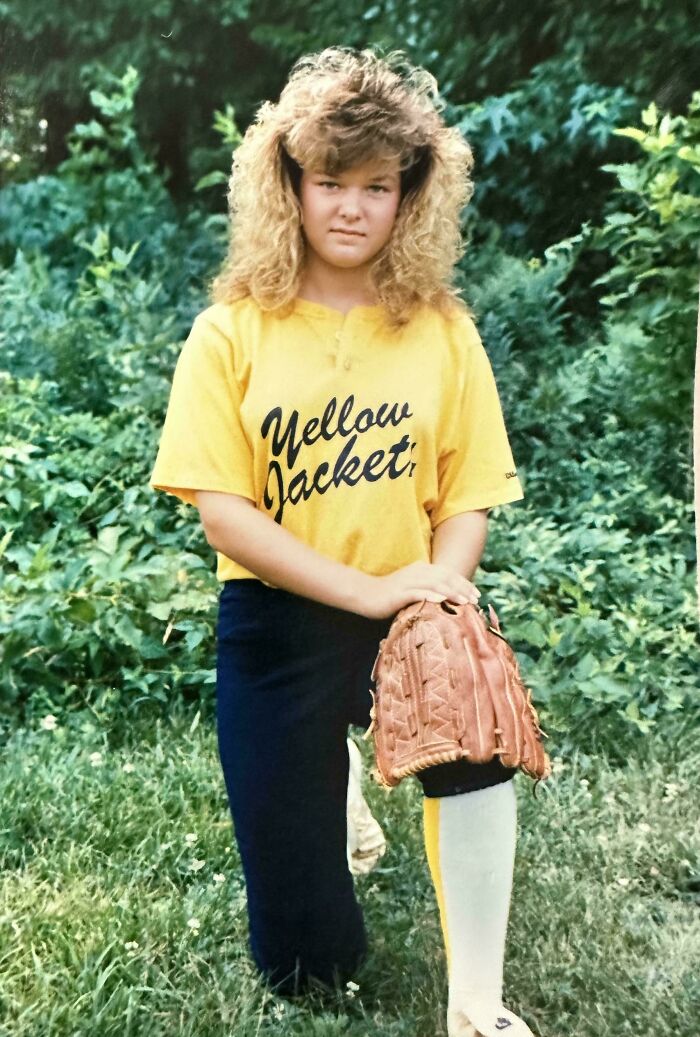 Young girl with an iconic 80s hairdo wearing a Yellow Jackets softball uniform holding a baseball glove outdoors.