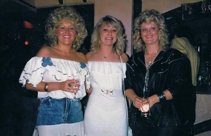 Three women posing at a party with iconic 80s hairdos featuring voluminous curls and teased styles.