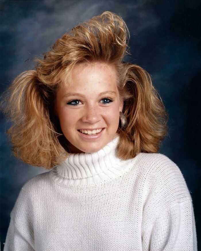 Teen girl with iconic 80s hairdo wearing a white turtleneck sweater smiling against a dark background