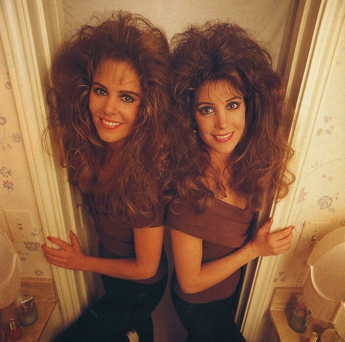 Two women with iconic 80s hairdos featuring voluminous teased curls, smiling in a vintage indoor setting.