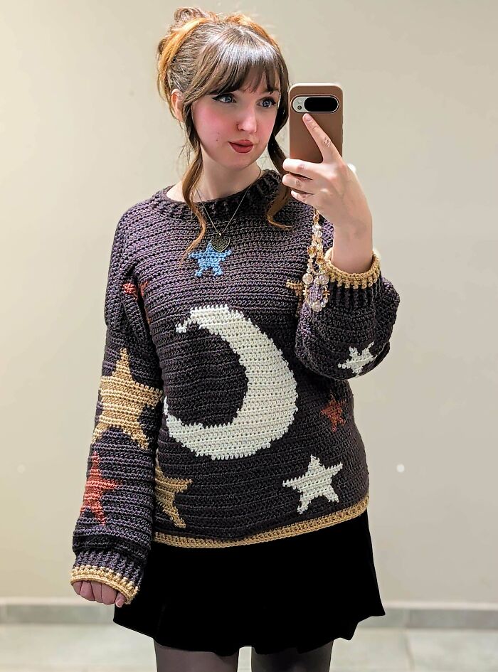 A young woman taking a selfie, showing off her awesome crocheted sweater with a moon and stars design.