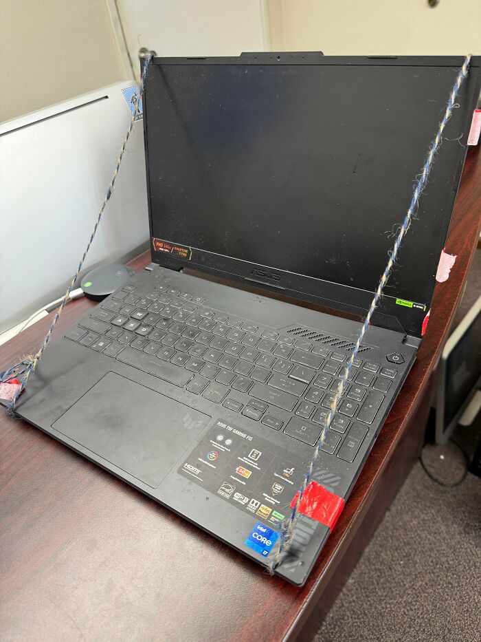 Laptop screen supported by makeshift string setup, showing a humorous tech support worker’s creative solution at a workstation.