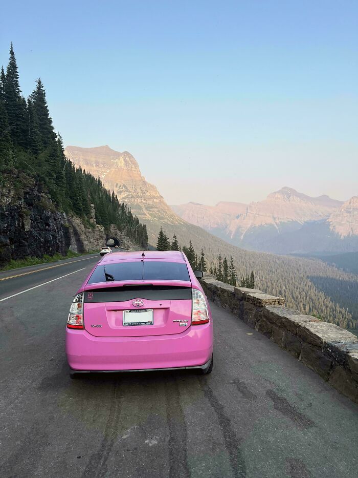 A pink car parked on a mountain road with evergreen trees and a vast mountain range in the background. Buy It For Life purchase.