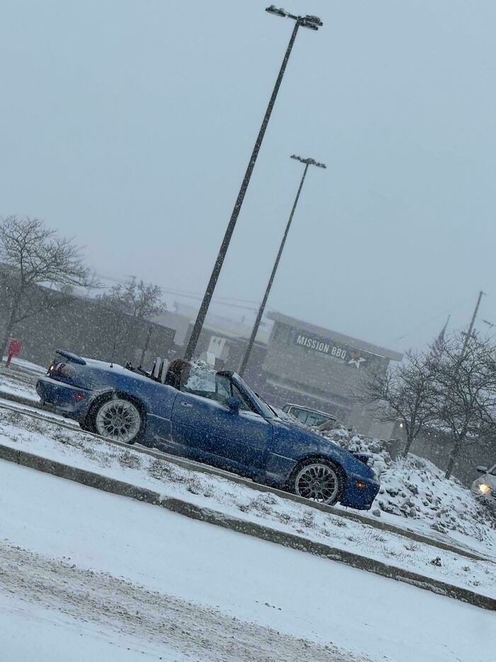 Blue convertible car parked in snowy weather with falling snowflakes under streetlights near a building and trees.