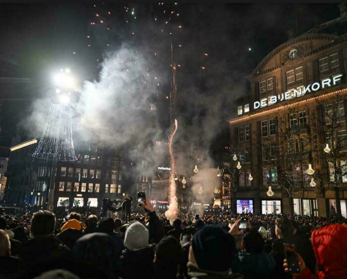 Crowd watching fireworks at night in a city square with smoke and bright lights, capturing a devil sunrise moment.