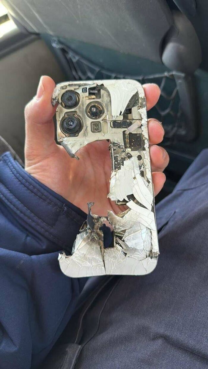 Hand holding a severely damaged smartphone with broken camera lenses and shattered back, illustrating extreme destruction.