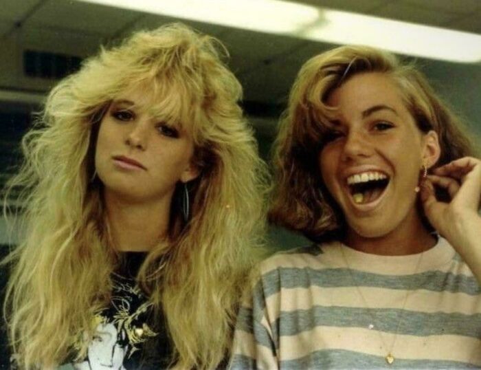 Two women with iconic 80s hairdos, one with voluminous teased hair and the other with natural curls, in a casual setting