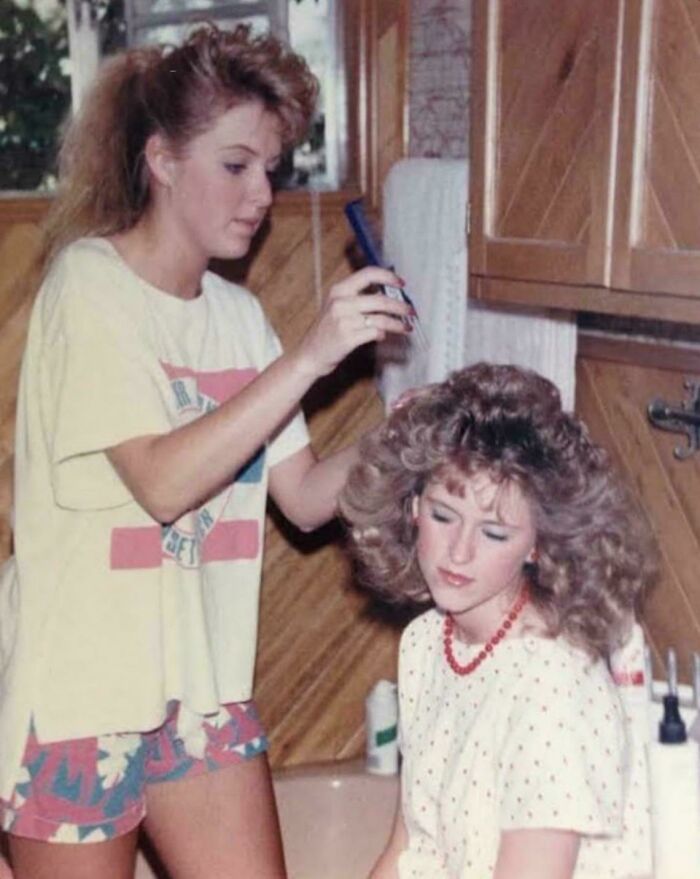 Woman styling another's voluminous iconic 80s hairdo in a vintage wooden-paneled bathroom setting.