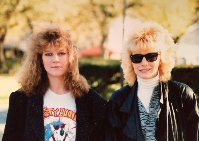 Two women sporting iconic 80s hairdos with voluminous curls and teased styles outdoors in casual vintage clothing.