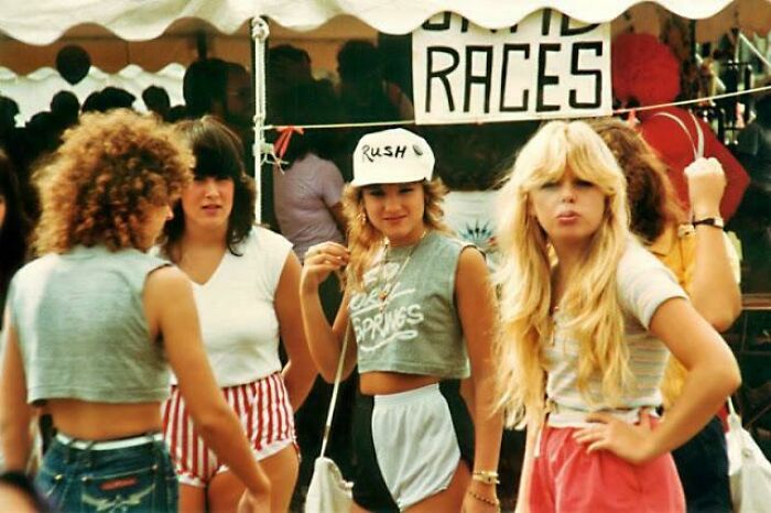 Four young women with iconic 80s hairdos in casual summer outfits standing near a tent at an outdoor event.