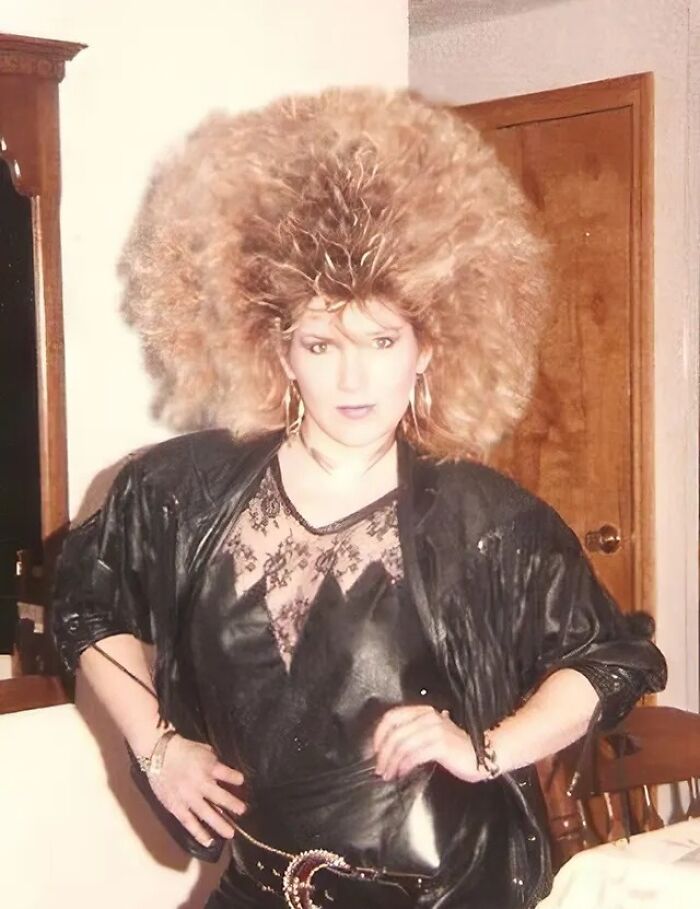 Woman with voluminous teased 80s hairdo wearing a black leather outfit and lace top indoors with hands on hips.