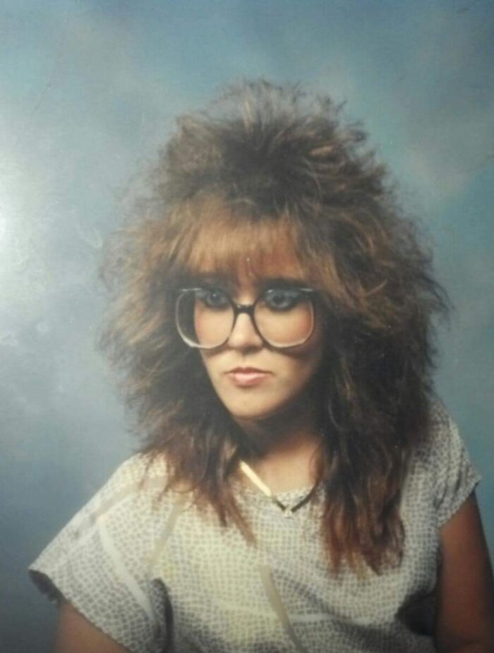Woman with iconic 80s hairdo and large glasses posing against a plain blue background in vintage style portrait.