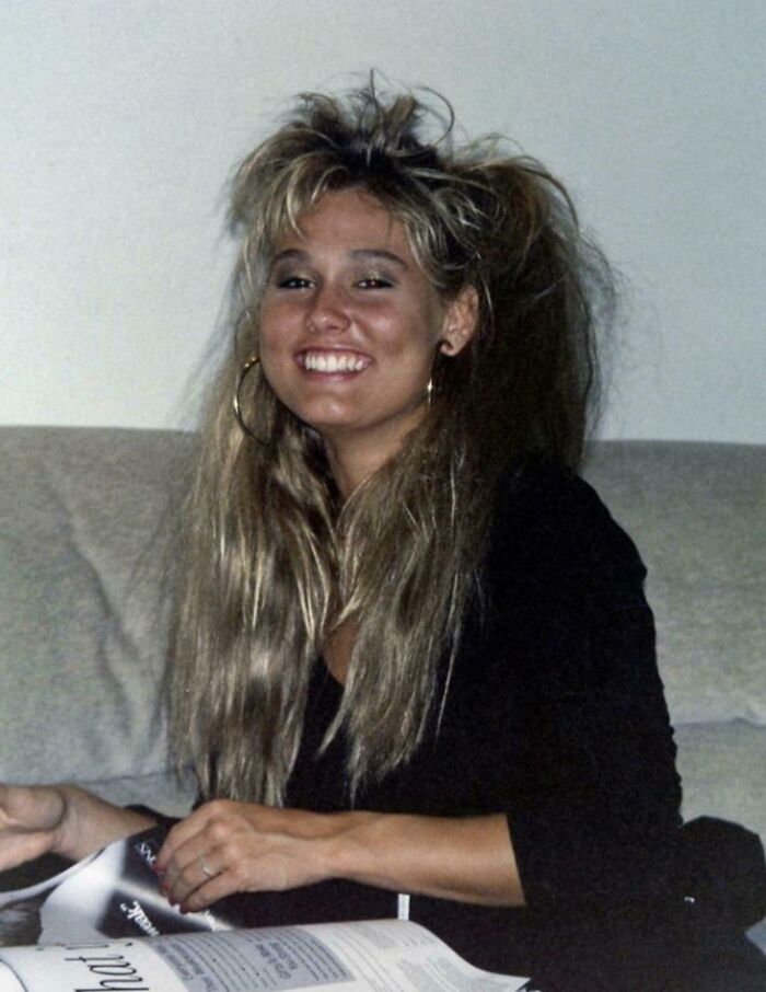 Young woman with an iconic 80s hairdo featuring voluminous, teased hair and large hoop earrings, smiling indoors.