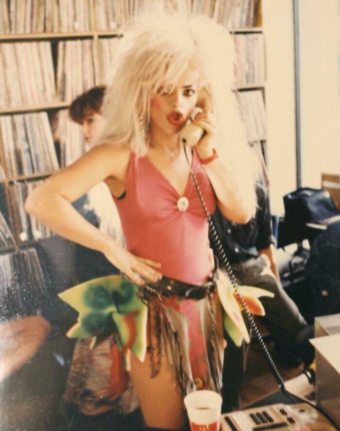 Woman with iconic 80s hairdos wearing a colorful outfit and talking on a corded phone in a vintage room with records.