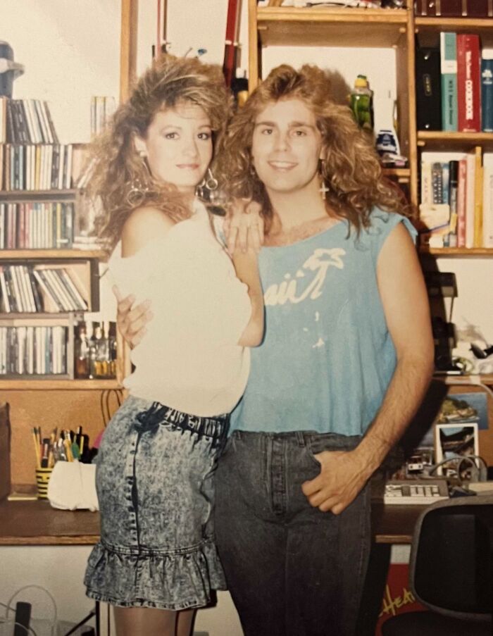A man and woman with iconic 80s hairdos pose together in a room filled with books and CDs, showcasing classic 80s hairstyles.