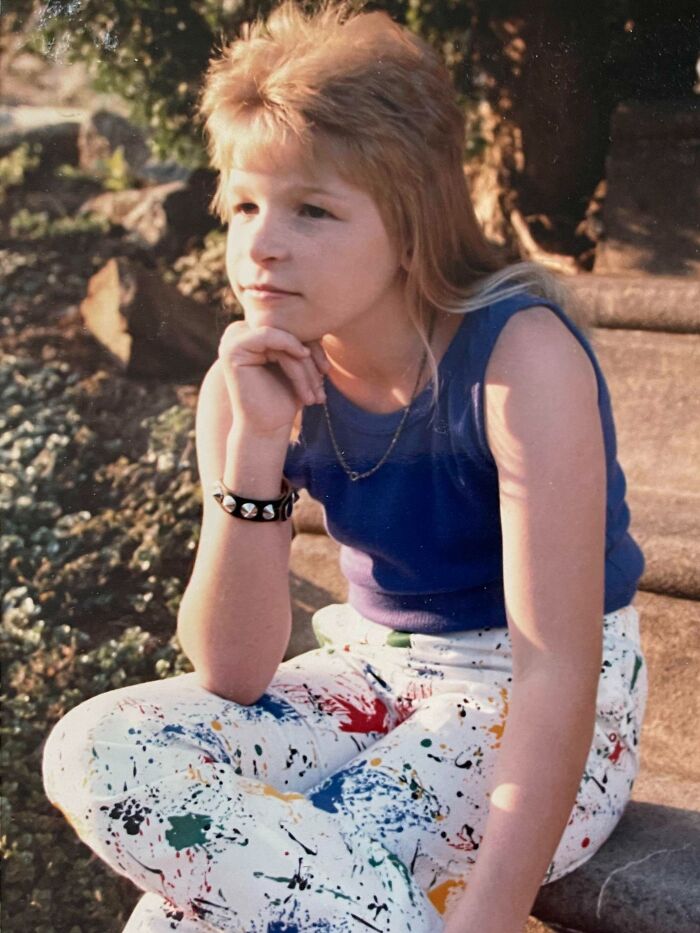 Young person with an iconic 80s hairdo wearing a blue tank top and paint-splattered pants sitting outdoors in sunlight.