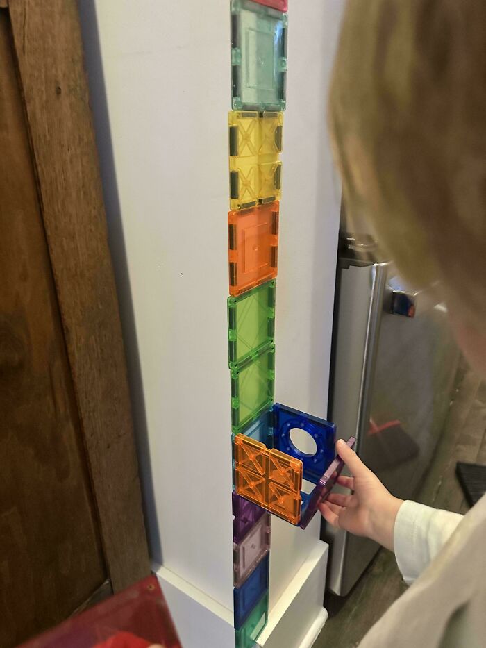 A goofy photo of a great dad's child playing with magnetic tiles, constructing a tall, colorful tower.