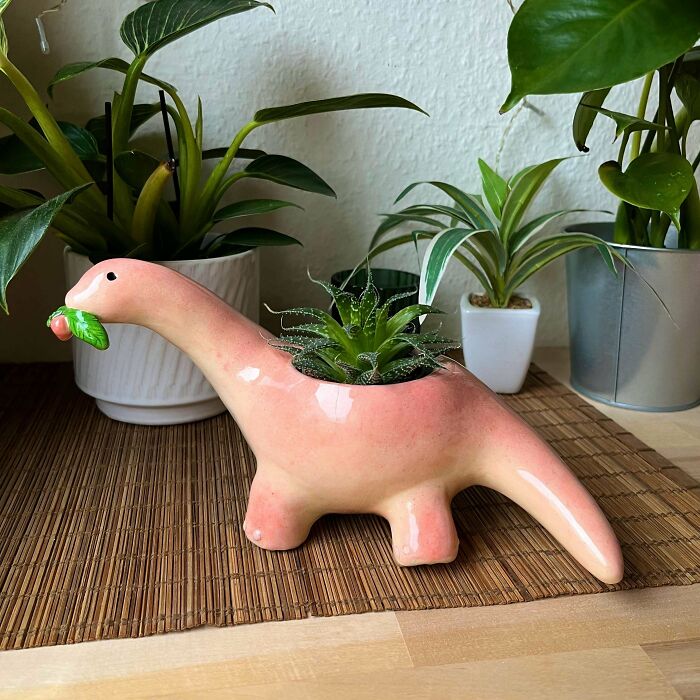 Pink dinosaur-shaped planter with a green succulent inside, surrounded by potted plants, showcasing a creative DIY win.