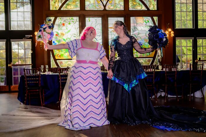 Two brides, one in a white crocheted dress and the other in a black gown, hold hands, looking at each other, happy.