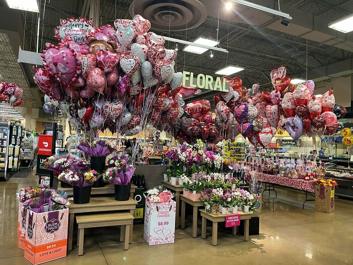 A supermarket floral section adorned with a large display of balloons and flowers, highlighting consumerism and price differences.