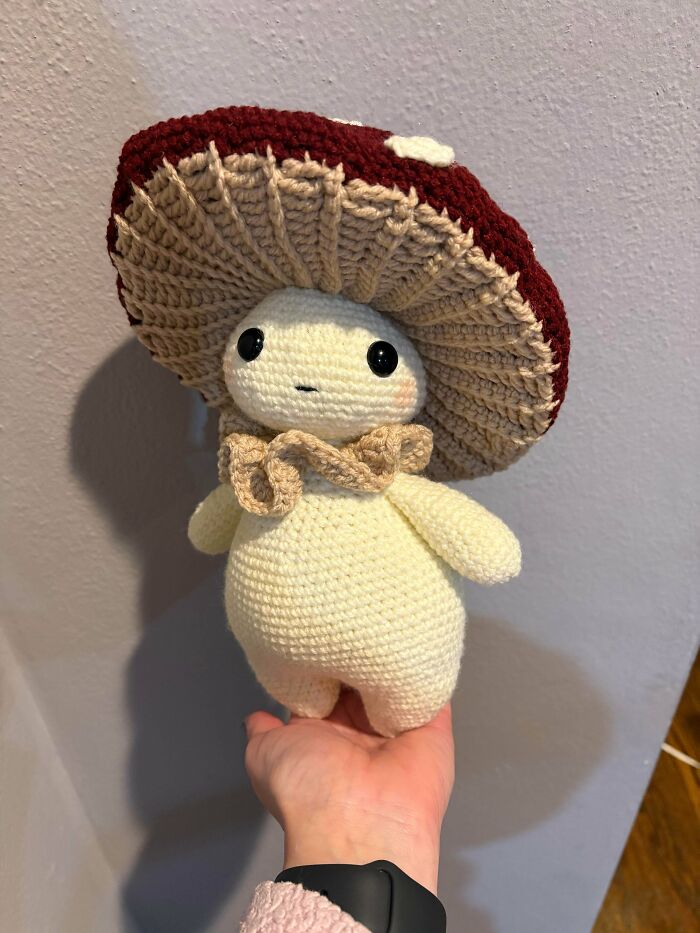 A hand holds a cute crocheted mushroom person with a red cap and beige ruffled collar. Crocheted awesome design.