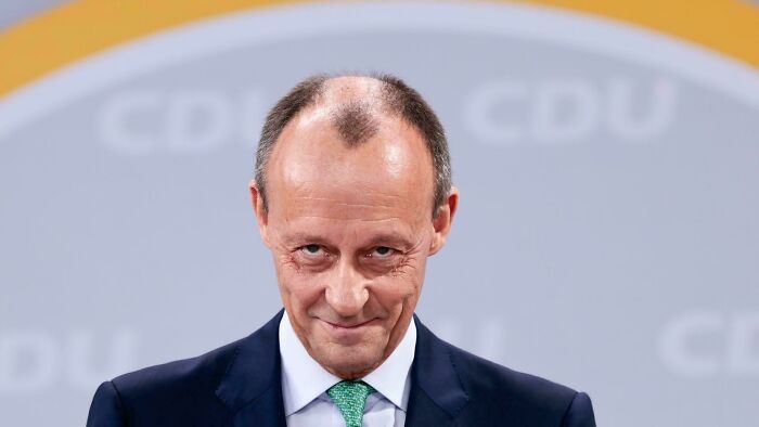 A man with a receding hairline and an unfortunate haircut, wearing a suit and smiling subtly.
