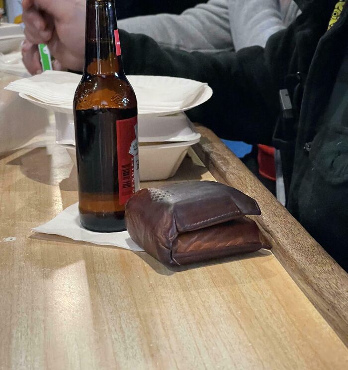 A large wallet placed next to a beer bottle on a wooden bar counter showcasing an absolute unit size comparison.