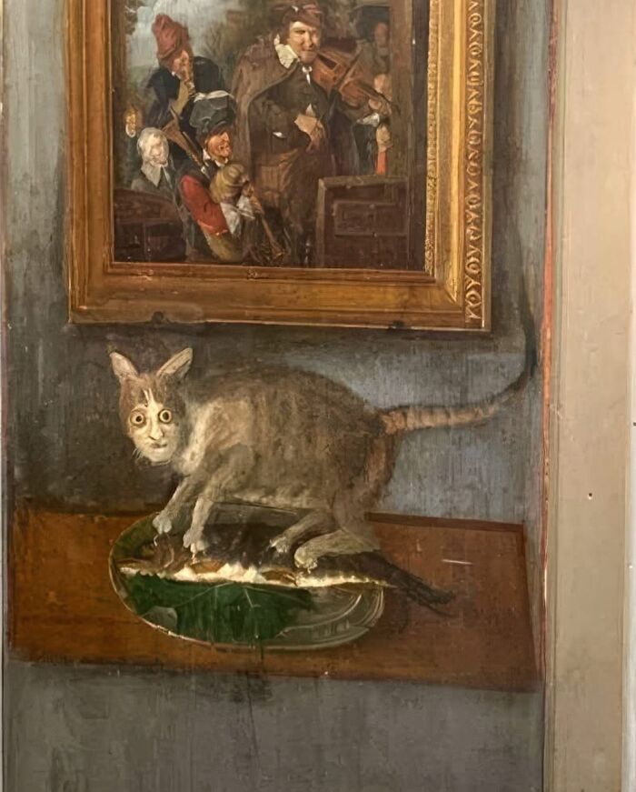 Surreal cat-themed artwork depicting a cat with a human-like face standing on a tray with fish beneath a framed painting.