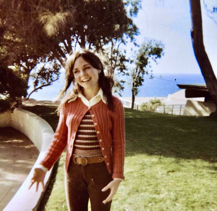 A candid old photo of a young woman with pigtails smiling outdoors, with trees and the ocean in the background, revealing what the world looked like generations ago.