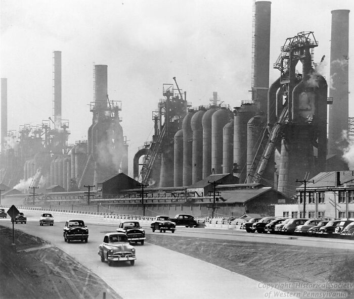 Black and white photo of old industrial buildings with smokestacks and vintage cars on a road nearby.