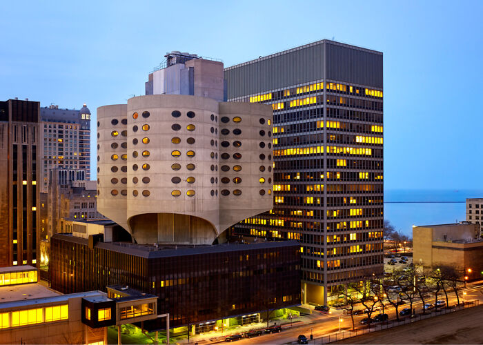 Unique interesting building with circular windows stands among modern city skyscrapers at dusk.