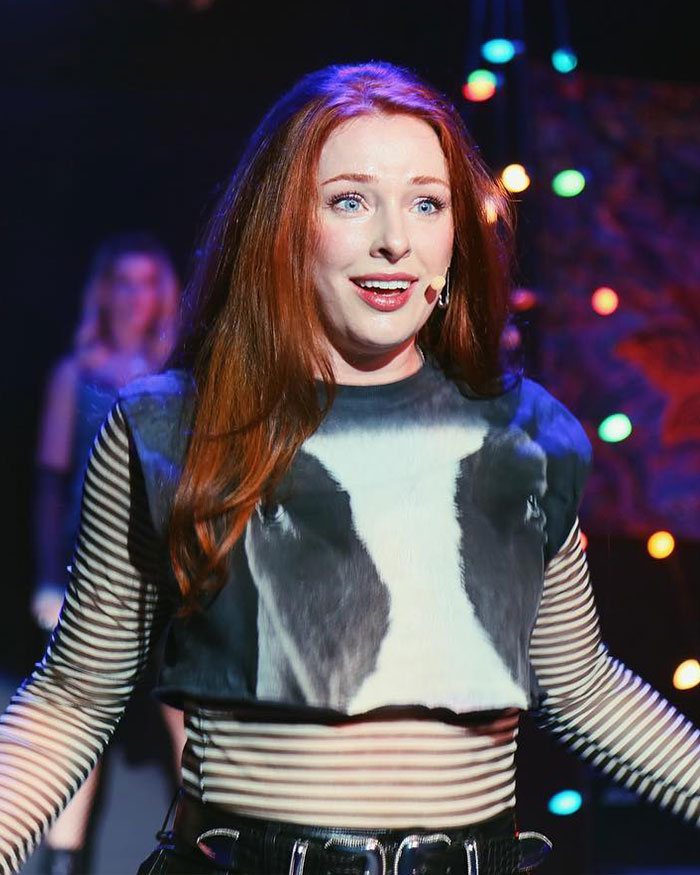 Woman with red hair wearing striped shirt and black top, performing on stage while sharing truth behind controversial firing.