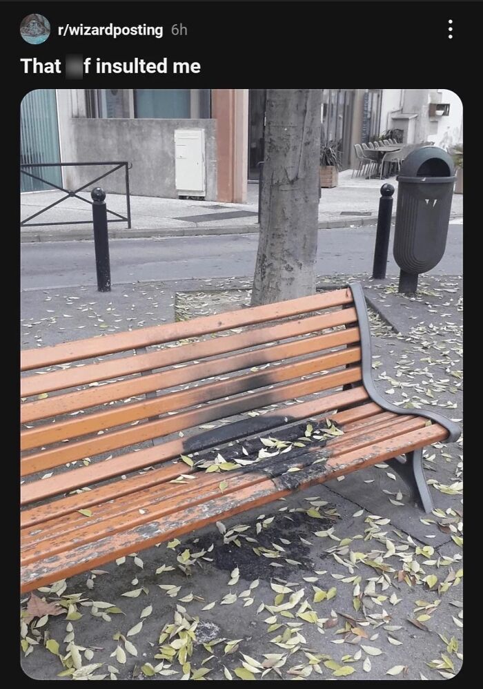 Wooden bench in park with burnt and damaged seat surrounded by fallen leaves, reflecting cynical humor meme style.