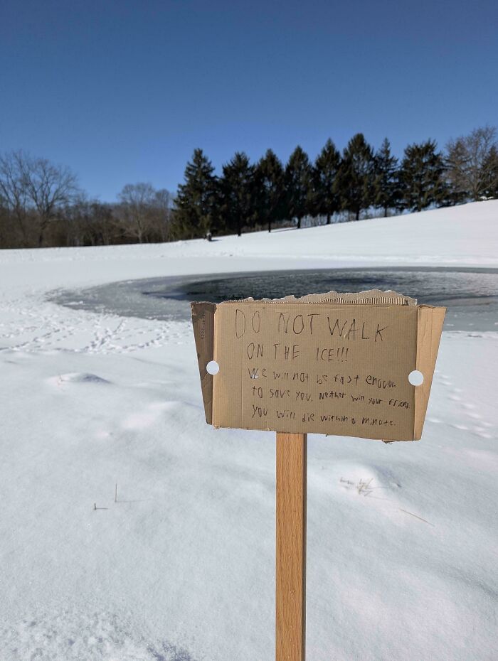 Handwritten warning sign on cardboard warns not to walk on the ice near a frozen pond in a snowy area.