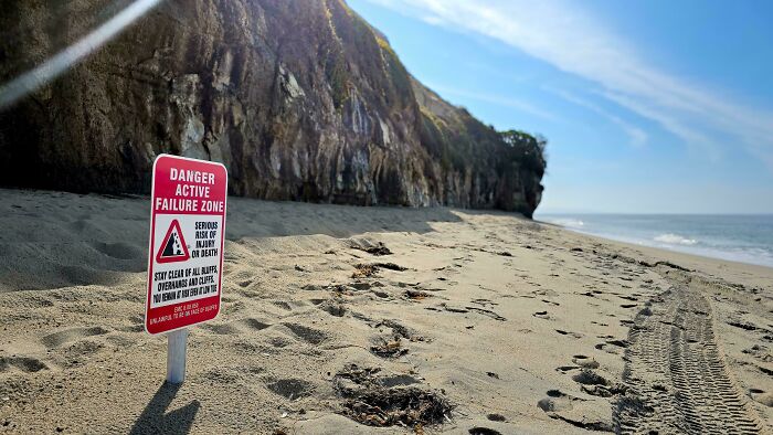 Warning sign on a sandy beach indicating an active failure zone with serious injury or death risk near cliffs and ocean.