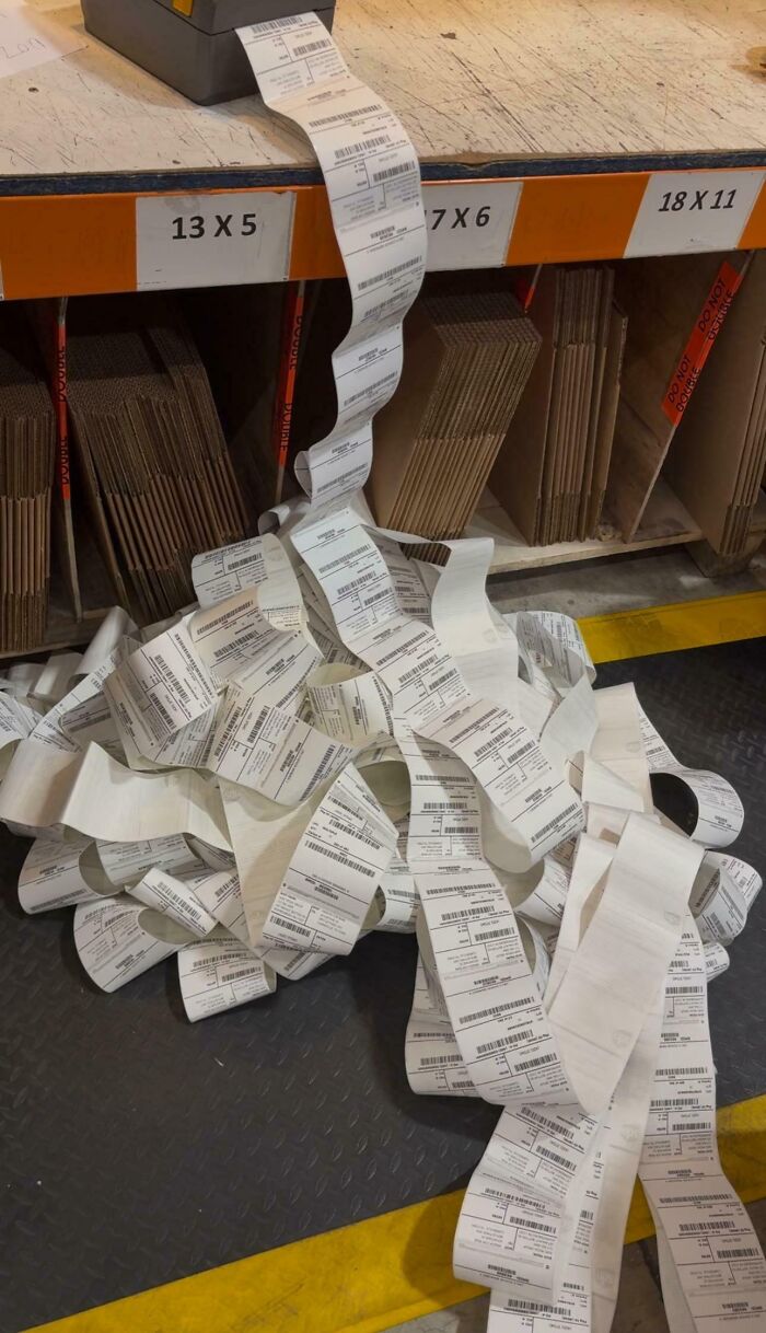 Long tangled receipt labels spilling on warehouse floor near cardboard box storage, showing tech support work frustration.