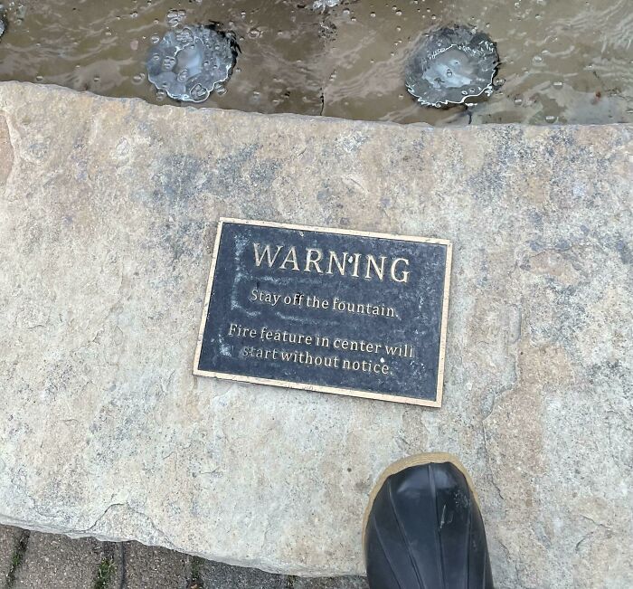 Warning sign on stone near water fountain indicating a fire feature may start without notice, one of the scariest signs spotted.