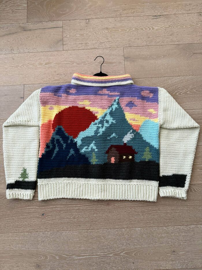 A stunning crocheted sweater with a vibrant mountain sunset scene and a cozy cabin. An awesome crocheted piece.