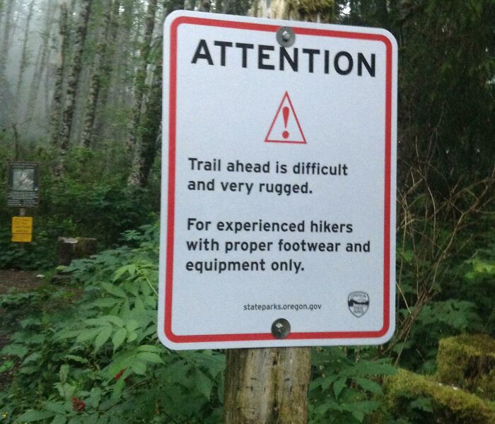 Warning sign in forest stating trail ahead is difficult and rugged for experienced hikers with proper footwear and equipment only.