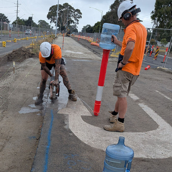 Two construction workers using a water jug attached to a traffic cone in a redneck engineering setup.