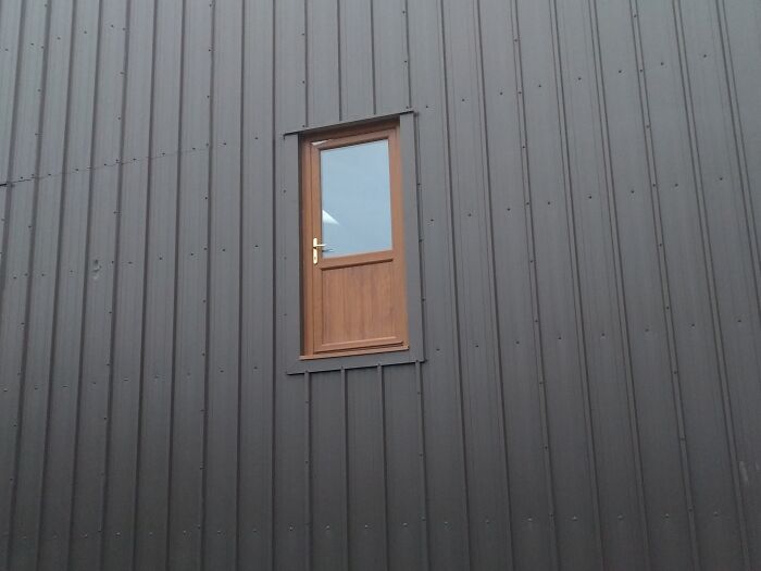 Small wooden door installed high on a black metal wall, an architectural fail with a door that leads nowhere.