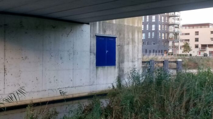 Blue door on a concrete wall under a bridge, part of architectural fail showing doors that lead absolutely nowhere.