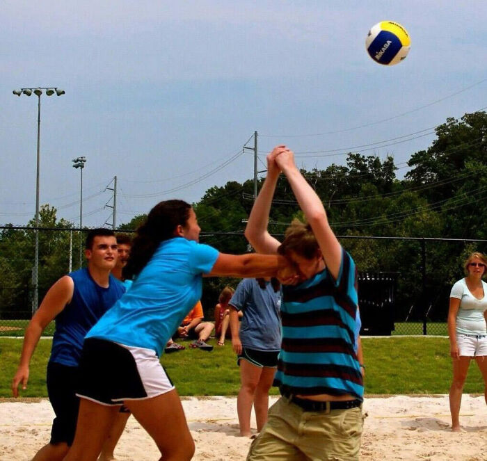 Two people accidentally collide while playing beach volleyball with others watching in a funny photos moment.