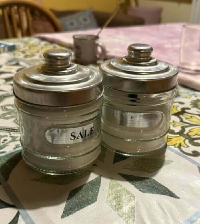 Two glass containers with metal lids on a table, showing a frustrating habit of incorrectly labeled salt and sugar jars.